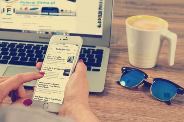 Mains tenant un smartphone affichant un article de presse, ordinateur portable en arrière-plan